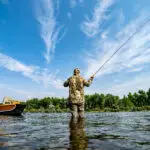 the back of a man fly fishing in a river, with drift boat next to him