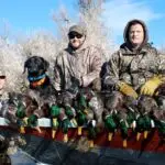 three men and a black dog showing off their duck hunt results