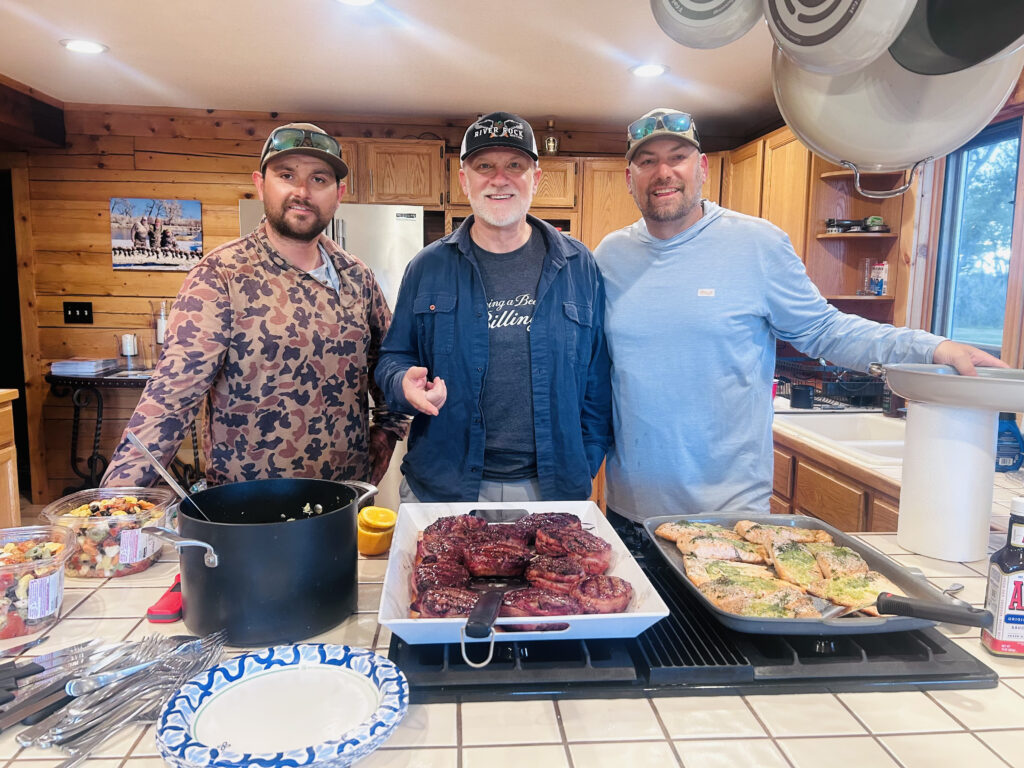 three men standing in a kitchen around a prepared dinner meal